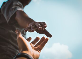 Dynamische Aufnahme aus der Untersicht zweier Hände, die sich gegenseitig zu greifen. Eine Hand streckt sich von unten nach oben, während die andere sich helfend entgegenstreckt, symbolisch für Unterstützung und Zusammenhalt, vor einem klaren blauen Himmel