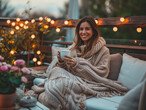 ine junge Frau sitzt lächelnd auf einer Terrasse, eingehüllt in eine große, gestrickte Decke, während sie eine Tasse hält. Die Terrasse ist mit Lichterketten beleuchtet und mit verschiedenen Pflanzen und Blumen dekoriert, die eine gemütliche und entspannte Atmosphäre schaffen.